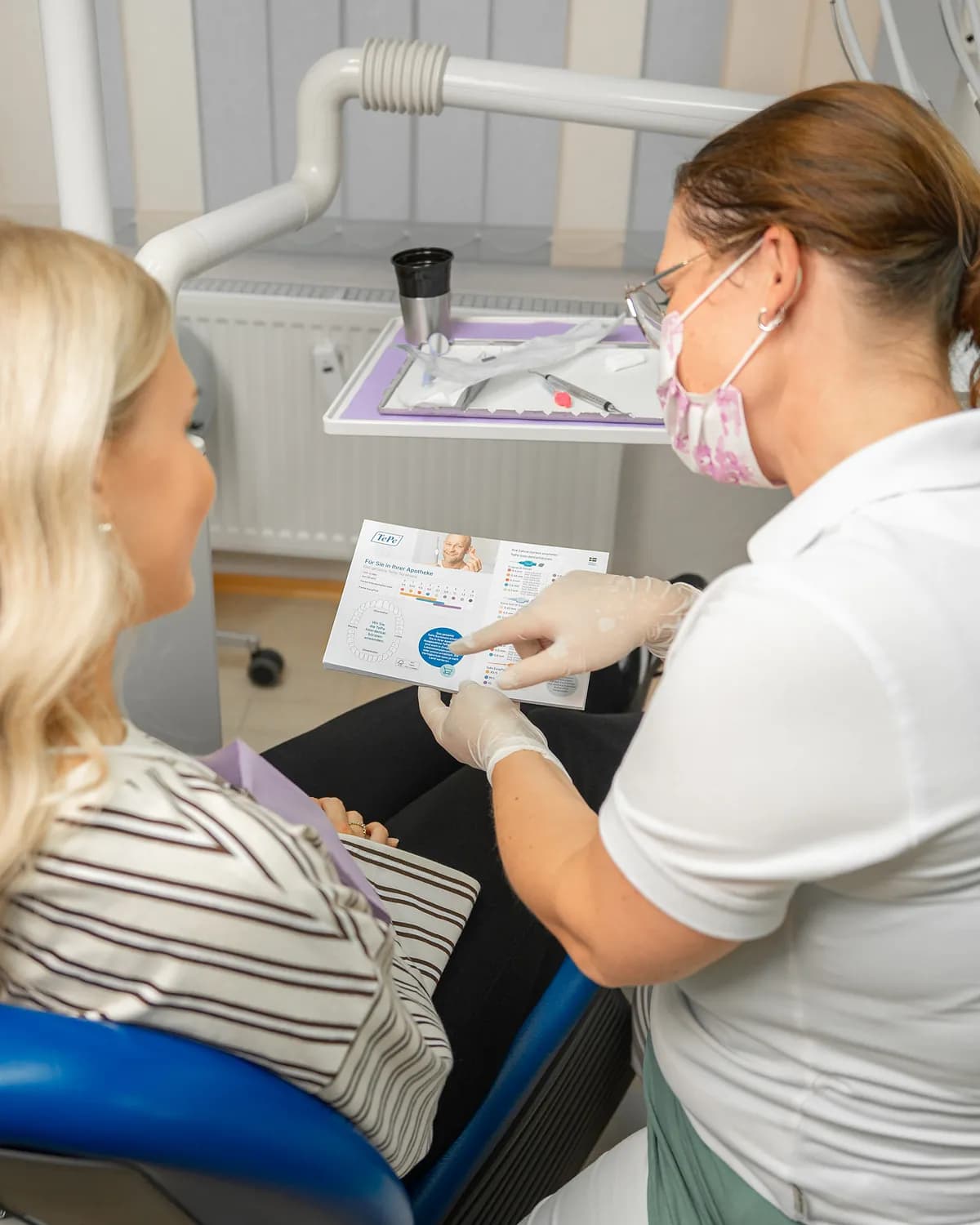 Mitarbeiterin erklärt einer Patientin im Behandlungsstuhl die Zahnpflege anhand einer Broschüre