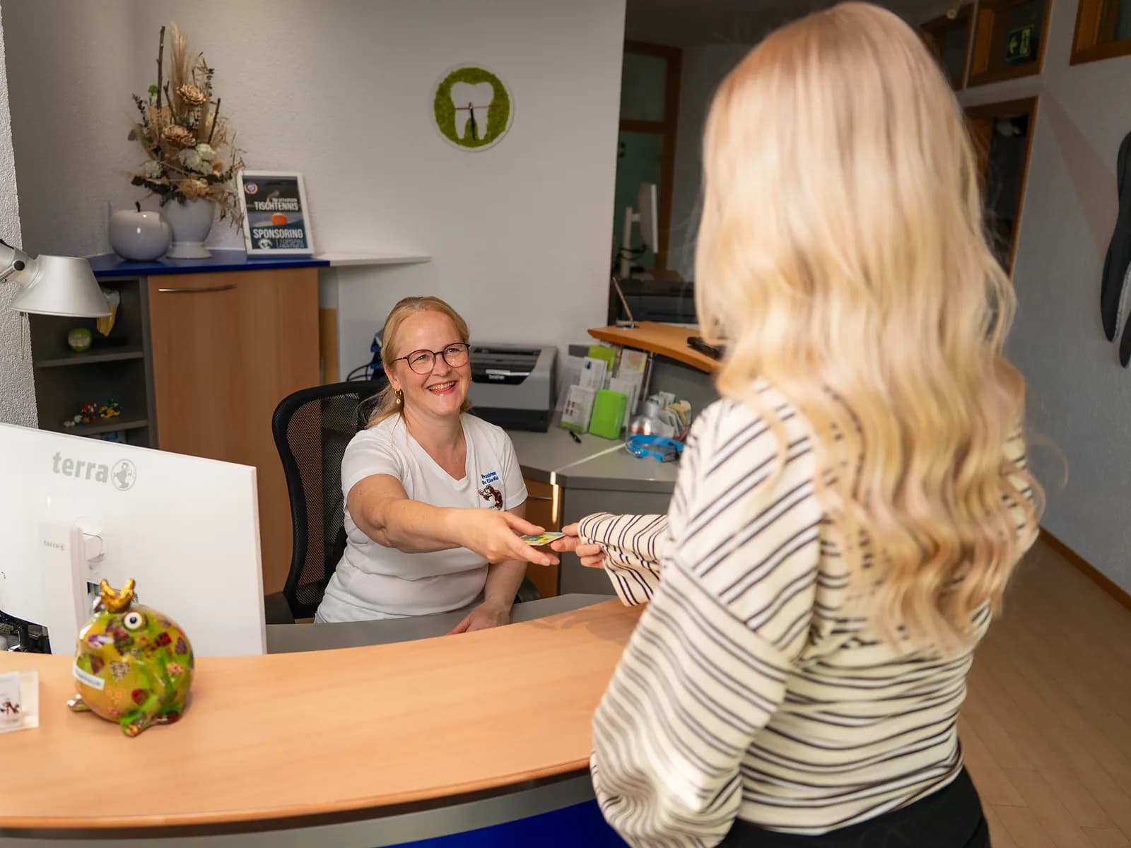 Mitarbeiterin am Empfang reicht einer Patientin ihre Unterlagen